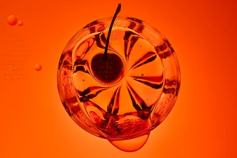 Overhead view of a glass with a cherry garnish, filled with an orange-red liquid. The light creates abstract reflections and patterns, with a few bubbles visible nearby on an orange background.
