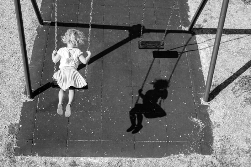 A young child with curly hair, seen from behind, swings on a playground swing. The child’s shadow is cast clearly on the ground tiles beneath them. The scene is in black and white.