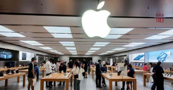 Inside an Apple Store, people browse products displayed on wooden tables. Large Apple logo is illuminated on the ceiling. Shelves filled with boxes and screens line the walls, and a busy atmosphere prevails with shoppers and staff interacting.
