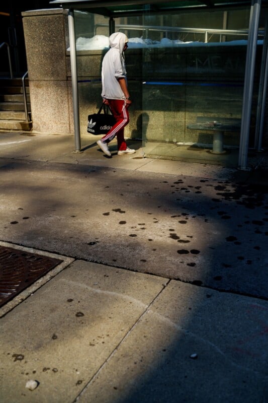 A person in a white hooded jacket and red pants walks past a glass structure. They carry an Adidas bag. Sunlight and shadows create patterns on the sidewalk.