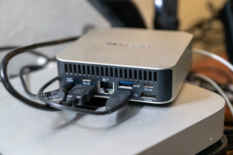 A compact silver computer hub with multiple connected cables sits on a desk. The hub features various ports such as HDMI, USB, and Ethernet, and is surrounded by a cluttered setup of wires and other electronic devices.