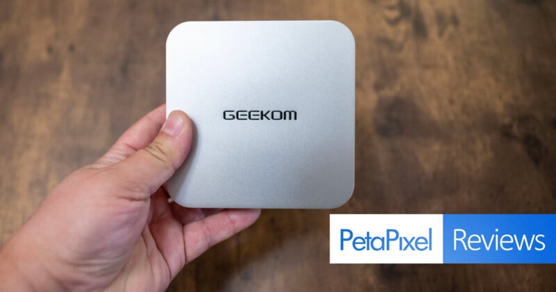 Hand holding a compact silver device with the brand name "GEEKOM" on top. The background is a wooden surface, and the "PetaPixel Reviews" logo is visible at the bottom right.