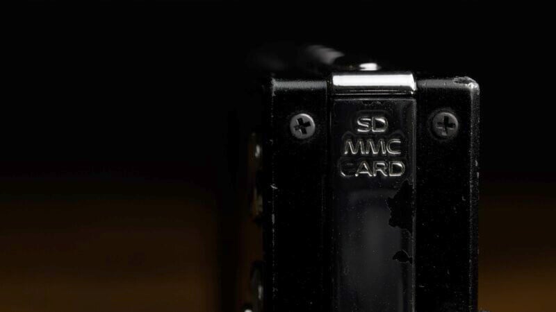Close-up of an SD/MMC card slot on a dark background with subtle lighting highlighting the metal screws and engravings. The slot is part of a black electronic device, and the surroundings are largely in shadow.