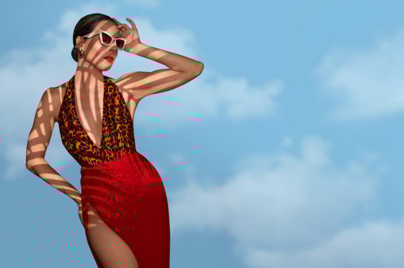 A woman in a red dress with a leopard-print top poses confidently against a blue sky with clouds. She wears white sunglasses, red lipstick, and casts striped shadows across her body.