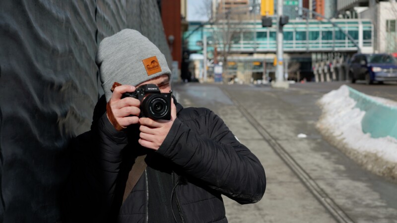 A person wearing a gray beanie and black jacket is holding a camera up to their face, taking a photo. They are standing on a city street with buildings and a few cars in the background. The street has patches of melting snow.