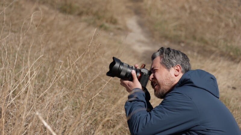 A person with gray hair and a beard is crouching in tall, dry grass, holding a camera to their face as they focus on taking a photo. They are wearing a blue jacket and appear deeply concentrated on their subject.